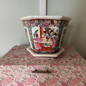 May include: A hexagonal ceramic planter with a matching saucer, featuring a white base decorated with colourful hand-painted scenes of figures in traditional attire, surrounded by intricate floral and geometric patterns in pink, green, and red.