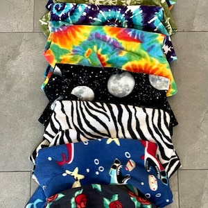May include: A stack of fleece fabric items with various patterns. Designs include black and white skulls, pink solid, green dinosaurs, tie-dye, zebra stripes, and a space theme with moons. The items are likely pet clothing.