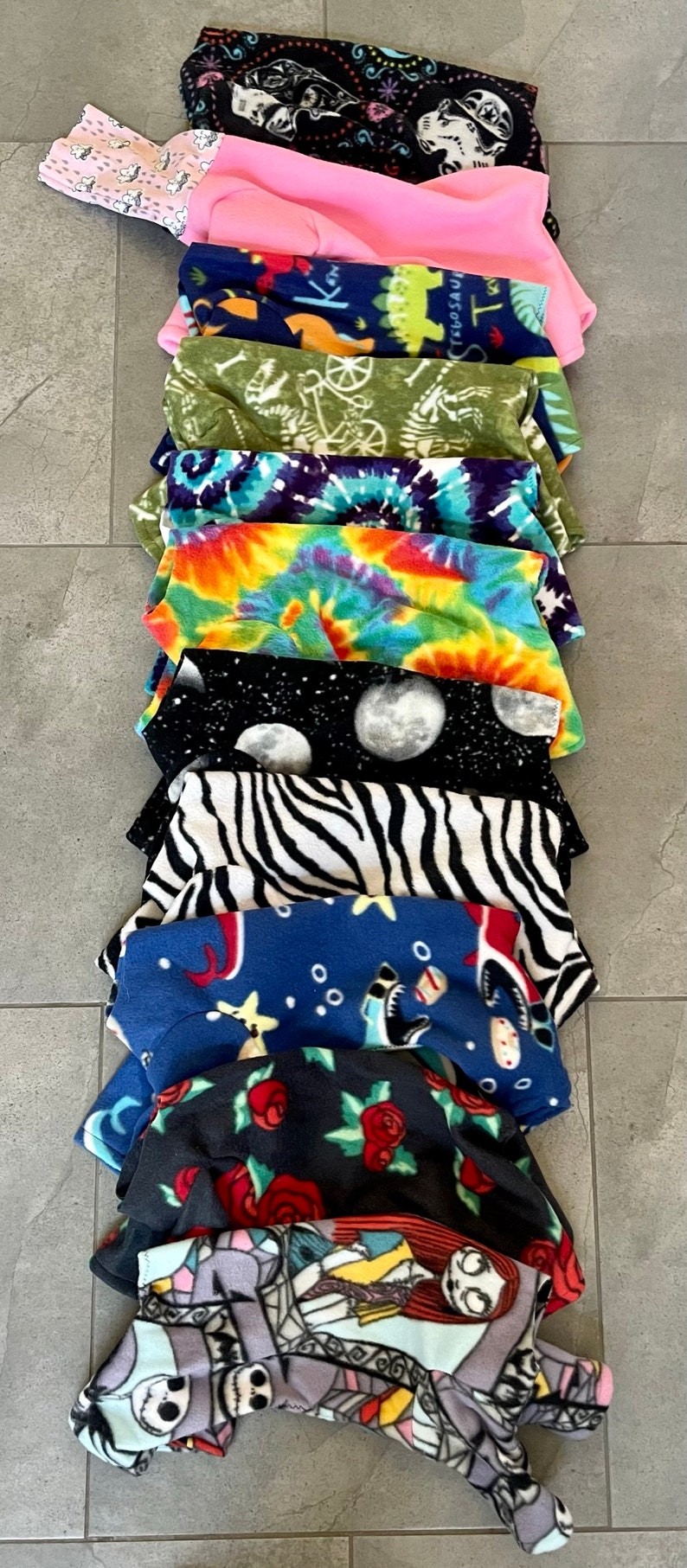 May include: A stack of fleece fabric items with various patterns. Designs include black and white skulls, pink solid, green dinosaurs, tie-dye, zebra stripes, and a space theme with moons. The items are likely pet clothing.