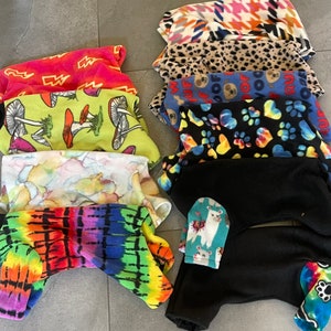 May include: A collection of fleece pet pajamas in various patterns and colors. Designs include tie-dye, mushrooms, paw prints, llamas, and houndstooth. The pajamas are designed to cover the pet's body, including the legs.