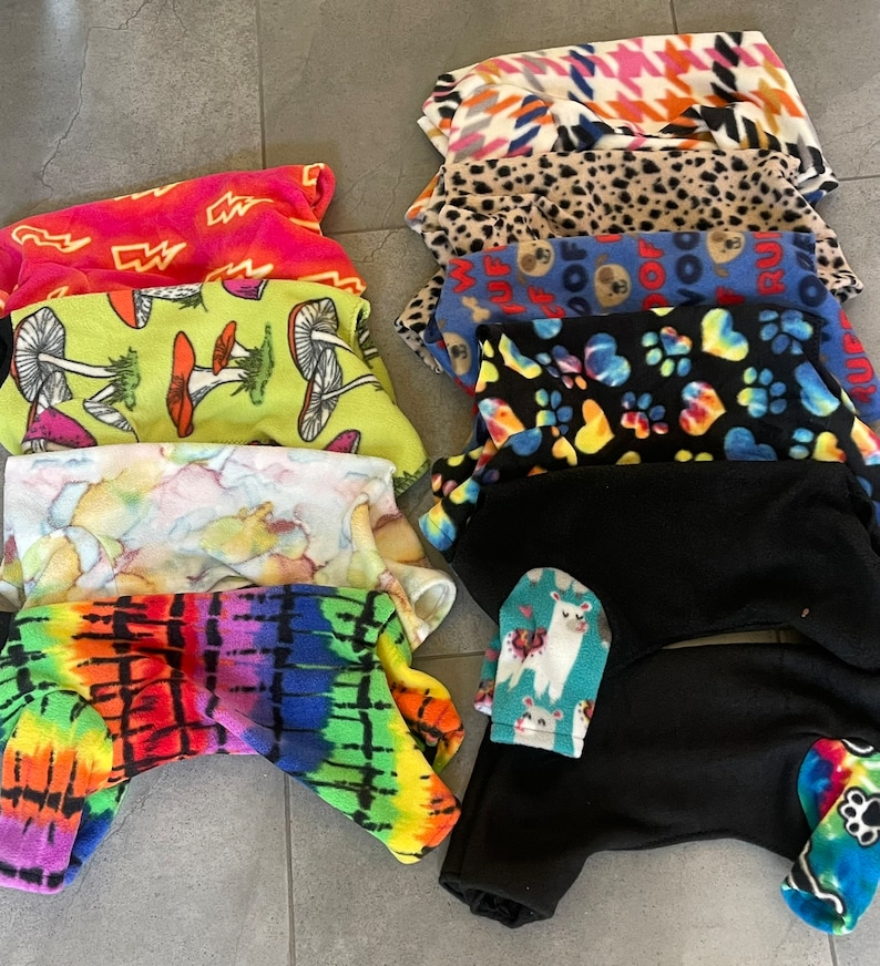 May include: A collection of fleece pet pajamas in various patterns and colors. Designs include tie-dye, mushrooms, paw prints, llamas, and houndstooth. The pajamas are designed to cover the pet's body, including the legs.