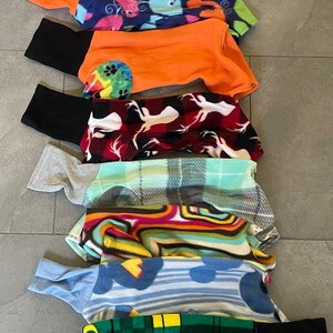 May include: A stack of dog clothes in various patterns and colors. The garments include a purple tie-dye, a blue whale print, an orange top with black sleeves, a red and white deer print, and a green plaid with the text "OREGON DUCKS".