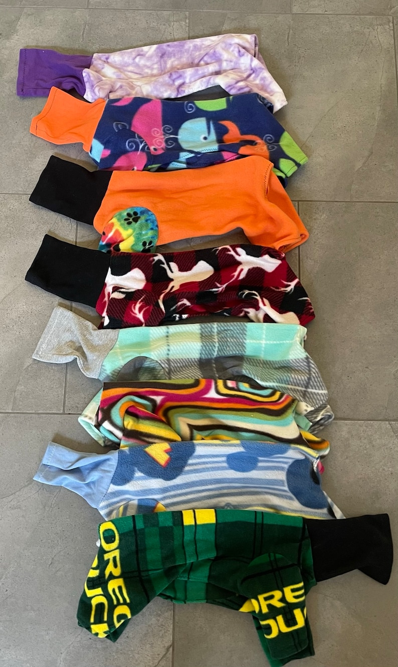 May include: A stack of dog clothes in various patterns and colors. The garments include a purple tie-dye, a blue whale print, an orange top with black sleeves, a red and white deer print, and a green plaid with the text "OREGON DUCKS".