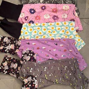 Könnte beinhalten: Eine Auswahl an Tierkleidung mit verschiedenen Mustern. Die Muster umfassen Weltraum-Designs, Donuts, Spiegeleier, kleine Figuren und eulenartige Totenköpfe. Die Farben sind Schwarz, Rosa, Blau, Lila und Grau.