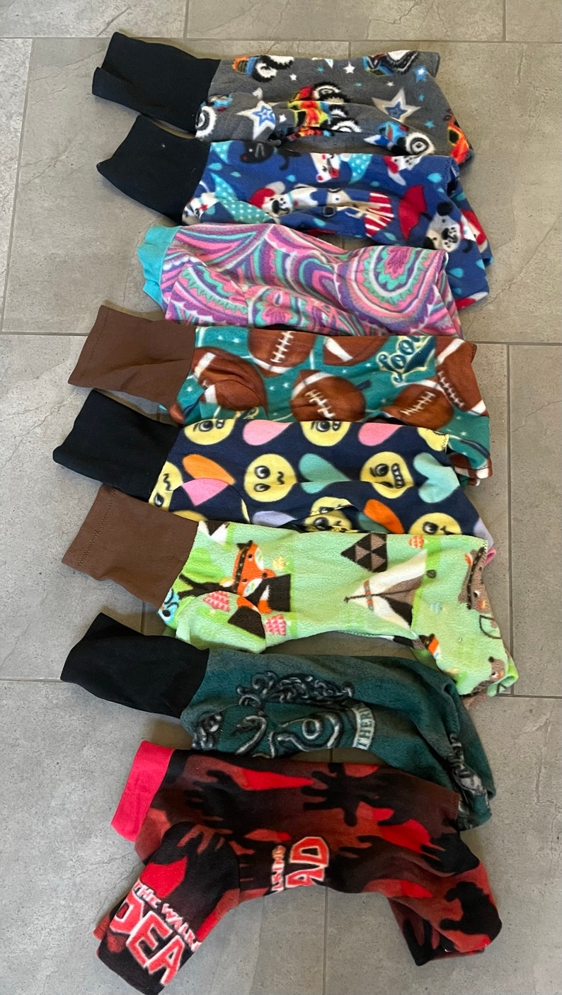 May include: A collection of pet pajamas in various patterns. Designs include stars, flags, paisley, footballs, smiley faces, and a zombie theme. Each pajama has a contrasting cuff at the ankle.