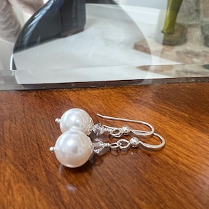 May include: A pair of elegant pearl earrings with silver hooks. Each earring features a large, round, white pearl and is accented with small, clear crystals. The earrings are displayed on a polished, dark wooden surface.