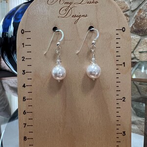 May include: A pair of pearl earrings with silver hooks and clear crystal accents. The earrings are displayed on a wooden stand with the text "Amy Lesho Designs". The stand has measurement markings.