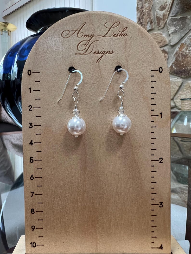 May include: A pair of pearl earrings with silver hooks and clear crystal accents. The earrings are displayed on a wooden stand with the text "Amy Lesho Designs". The stand has measurement markings.