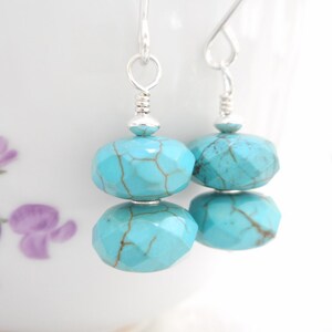 Puede incluir: Un par de pendientes de turquesa con ganchos de plata. Las piedras de turquesa son facetadas y tienen un color azul claro con vetas marrones.