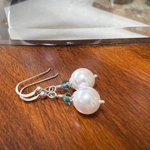 May include: A pair of elegant earrings featuring large, round, white faux pearls. The earrings have silver-colored hooks and are accented with small, teal-colored crystals. The jewelry rests on a dark wooden surface.