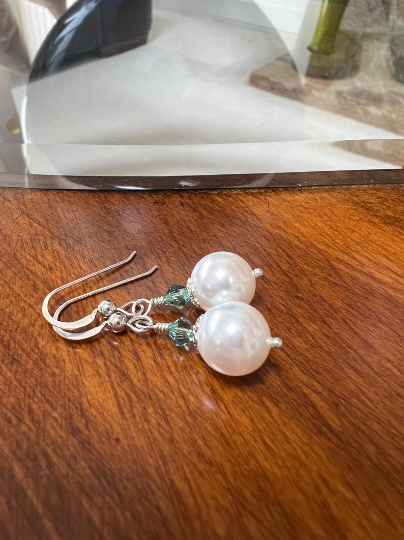 May include: A pair of elegant earrings featuring large, round, white faux pearls. The earrings have silver-colored hooks and are accented with small, teal-colored crystals. The jewelry rests on a dark wooden surface.