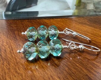 May include: A pair of drop earrings featuring three faceted, translucent light green glass beads on each silver wire. The beads are arranged in a vertical row, secured with silver headpins and attached to silver ear hooks with a small ball detail.
