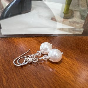 May include: A pair of elegant earrings featuring large, round, white faux pearls. The earrings have silver-colored hooks and delicate wirework with small, clear crystal accents. The earrings are displayed on a polished, dark wood surface.