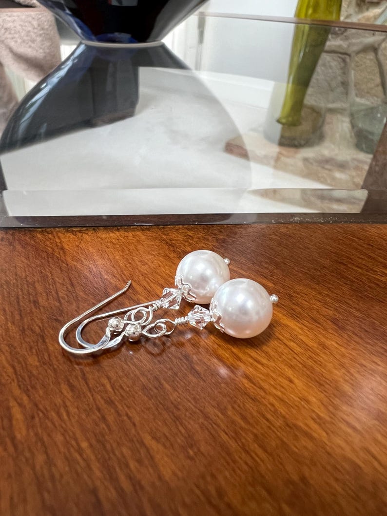 May include: A pair of elegant earrings featuring large, round, white faux pearls. The earrings have silver-colored hooks and delicate wirework with small, clear crystal accents. The earrings are displayed on a polished, dark wood surface.