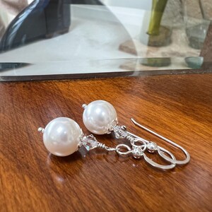 May include: A pair of elegant pearl earrings. Each earring features a large, round, white pearl suspended from a silver-toned hook. The hooks are adorned with small, clear crystals. The earrings are displayed on a dark wooden surface.