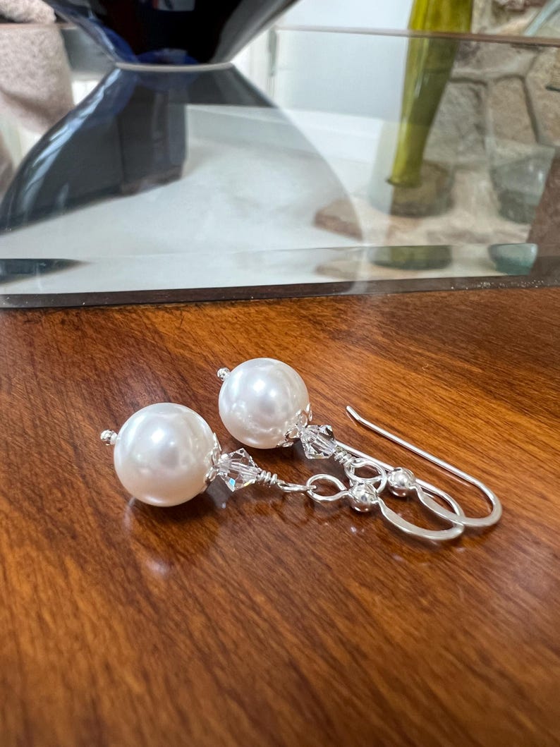 May include: A pair of elegant pearl earrings. Each earring features a large, round, white pearl suspended from a silver-toned hook. The hooks are adorned with small, clear crystals. The earrings are displayed on a dark wooden surface.