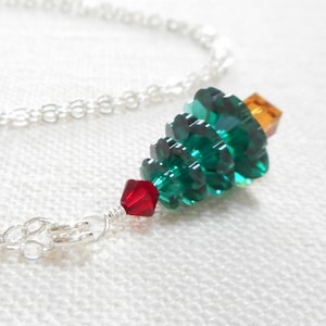 May include: A silver chain necklace with a green crystal Christmas tree pendant. The tree is decorated with a red crystal at the bottom and an orange crystal at the top.