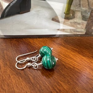 May include: A pair of dangle earrings featuring round, polished malachite beads with distinct dark and light green banded patterns. The beads are secured with silver-toned wire wrapping and attached to simple silver-toned shepherd hook ear wires.