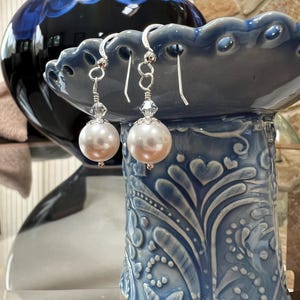 May include: Elegant pearl drop earrings with silver-tone hooks and crystal accents. The earrings feature large, round, off-white pearls. The earrings are displayed on a blue decorative ceramic bowl with a floral design.