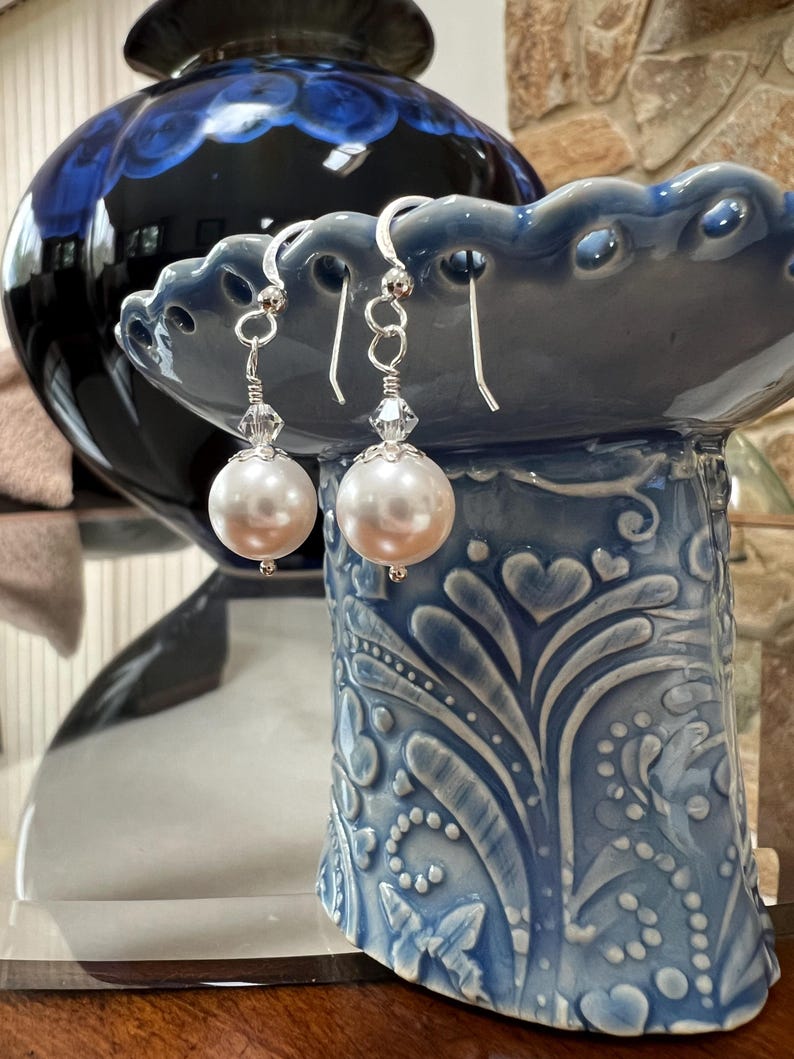 May include: Elegant pearl drop earrings with silver-tone hooks and crystal accents. The earrings feature large, round, off-white pearls. The earrings are displayed on a blue decorative ceramic bowl with a floral design.