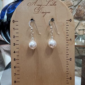 May include: A pair of pearl drop earrings displayed on a wooden stand. The earrings have silver hooks and delicate chains leading to large, round, white pearls. The stand is marked with measurements and the text "Amy Lesko Designs".