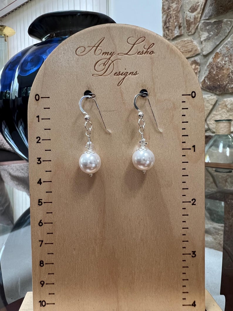 May include: A pair of pearl drop earrings displayed on a wooden stand. The earrings have silver hooks and delicate chains leading to large, round, white pearls. The stand is marked with measurements and the text "Amy Lesko Designs".