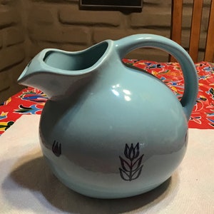 Puede incluir: Una jarra de cerámica azul claro con cuerpo redondeado y un pico ancho. La jarra tiene un diseño de tulipán en blanco y negro en la parte delantera.