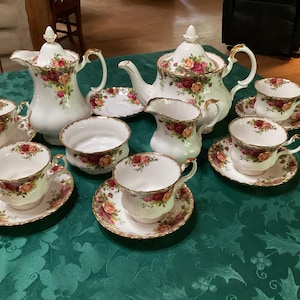 Puede incluir: Un juego de té completo de porcelana blanca con un diseño floral de rosas. El juego incluye una tetera, una jarra de leche, un azucarero, una fuente y varias tazas y platillos. Los bordes de cada pieza están adornados con detalles dorados. El juego está sobre un mantel verde.