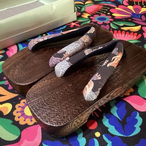 May include: A pair of traditional Japanese geta sandals with dark wooden platforms and patterned fabric straps. The straps feature floral and dragon designs in shades of gray, orange, and blue. The sandals rest on a colorful floral patterned fabric.