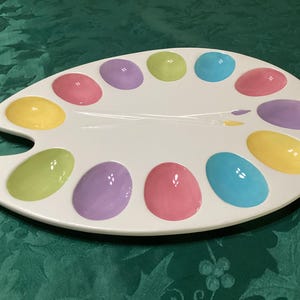 May include: A white ceramic artist palette serving tray featuring twelve oval-shaped indentations in various pastel colors including pink, purple, yellow, lime green, and light blue, with two painted paintbrushes crossed in the center.