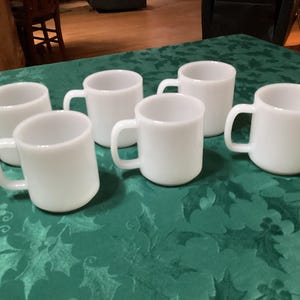 May include: Six white ceramic mugs with rounded bodies and attached handles are arranged on a green tablecloth. The mugs are evenly spaced and appear to be empty. The mugs are a solid, opaque white colour.