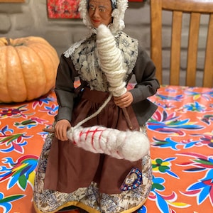 May include: A hand-crafted doll wearing a brown dress with a white lace collar and a white cap. The doll is holding a spindle with white yarn and is sitting on a colorful patterned tablecloth. The doll is labeled "Made in France".