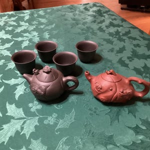 May include: A tea set featuring two teapots and four small cups. One teapot is dark brown with floral detailing, the other is reddish-brown with embossed patterns. The cups and teapots are arranged on a green tablecloth.