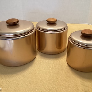 Puede incluir: Conjunto de tres botes de cobre con tapas de madera. Los botes son de color cobre claro y tienen un acabado brillante. Las tapas son de madera marrón oscura.