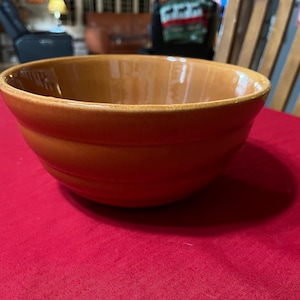 Può includere: Una grande ciotola rotonda in ceramica arancione-marrone. La ciotola ha un bordo largo e una superficie leggermente strutturata. Si trova su una superficie rossa. La ciotola viene probabilmente utilizzata per la preparazione di alimenti.