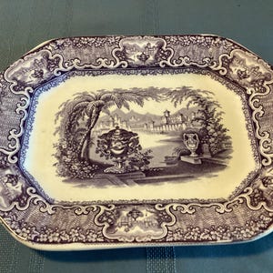 May include: An antique rectangular serving platter with a purple and white landscape design. The center features a detailed scene with palm trees, buildings, and decorative urns. The border has a floral and scroll pattern.