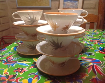 May include: A stack of four white teacups and saucers with a brown rim and a leaf design. The cups and saucers are stacked on a green tablecloth with a floral pattern.