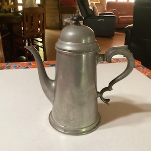 Op de afbeelding: Een zilveren koffiekan van tin met een zwarte knop op de deksel en een decoratieve handgreep. De kan staat op een wit oppervlak.