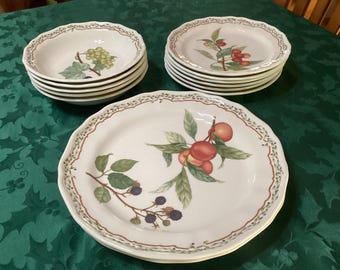 May include: A collection of white ceramic dishes with scalloped edges. The set includes bowls, plates, and a large platter, all decorated with fruit and leaf designs in shades of green, red, and brown. The dishes are arranged on a green tablecloth.