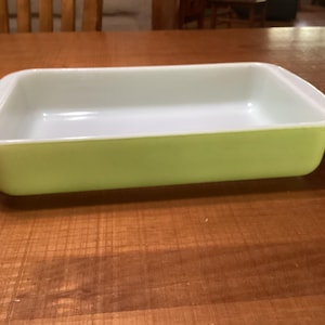 Puede incluir: Una fuente rectangular para hornear con exterior verde claro e interior blanco. La fuente tiene esquinas redondeadas y asas en cada extremo. La fuente está sobre una superficie de madera.