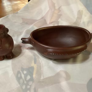 May include: Two terracotta-colored ceramic pieces: a small, rounded vase with a detailed top and a bowl with a fin-like handle. The bowl has a decorative rim. Both pieces are on a patterned surface.