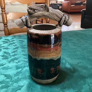 May include: A cylindrical ceramic vase featuring a glossy glaze with horizontal bands of black, deep red, and tan colours. The top and bottom rims have a textured cross-hatch pattern. A piece of weathered driftwood serves as a handle, secured by black ties.