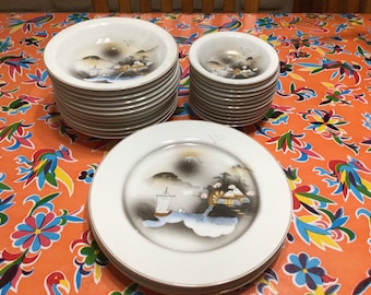 May include: A set of white ceramic plates and bowls with a Japanese-inspired design featuring a mountain, a boat, and a water wheel. The plates and bowls have a gold trim.