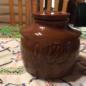 Op de afbeelding: Een bruine keramische pot met een deksel, op een tafelkleed met patroon. De pot heeft reliëfletters en een glanzende afwerking. De pot is een decoratief keukenartikel.