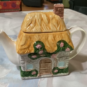 May include: A ceramic teapot shaped like a cottage. The roof is straw-colored, and the walls are stone-like with green vines and pink flowers. It has a chimney and a white handle and spout.