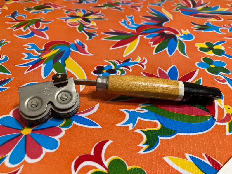 May include: A vintage can opener with a wooden handle and a silver metal cutting mechanism. The opener has two wheels with serrated edges for opening cans.