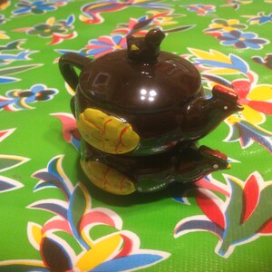 以下が含まれることがあります： 鶏のデザインが施された茶色の陶器製のティーポット。ティーポットには、小さな鶏が乗った蓋が付いています。ティーポットは黄色と赤のアクセントで飾られています。
