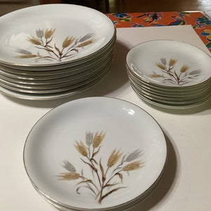 Può includere: Un set di piatti in ceramica bianca con un disegno di spighe di grano marrone, oro e grigio. I piatti hanno un bordo dorato e sono impilati in più pile. Sono visibili varie dimensioni di piatti.