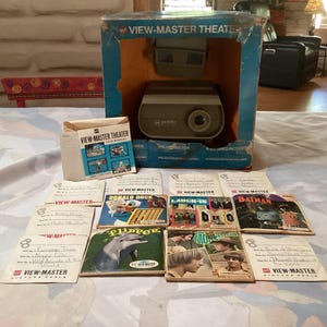 May include: A vintage View-Master Theater set with a projector and several View-Master reels. The box is blue and orange, with the words "View-Master Theater" printed on it. The reels feature images of Donald Duck, Batman, and Flipper.