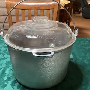 May include: A large, silver-colored cooking pot with a clear glass lid and a metal handle. The pot has a textured surface and is sitting on a green tablecloth. The lid has a small handle and a decorative design.